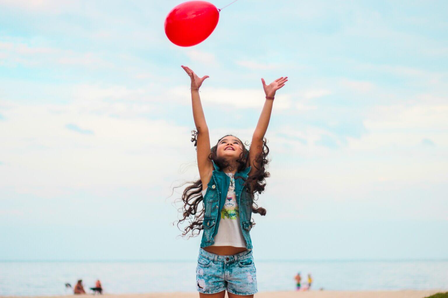 girl raising her hands reaching for a red balloon in the sky